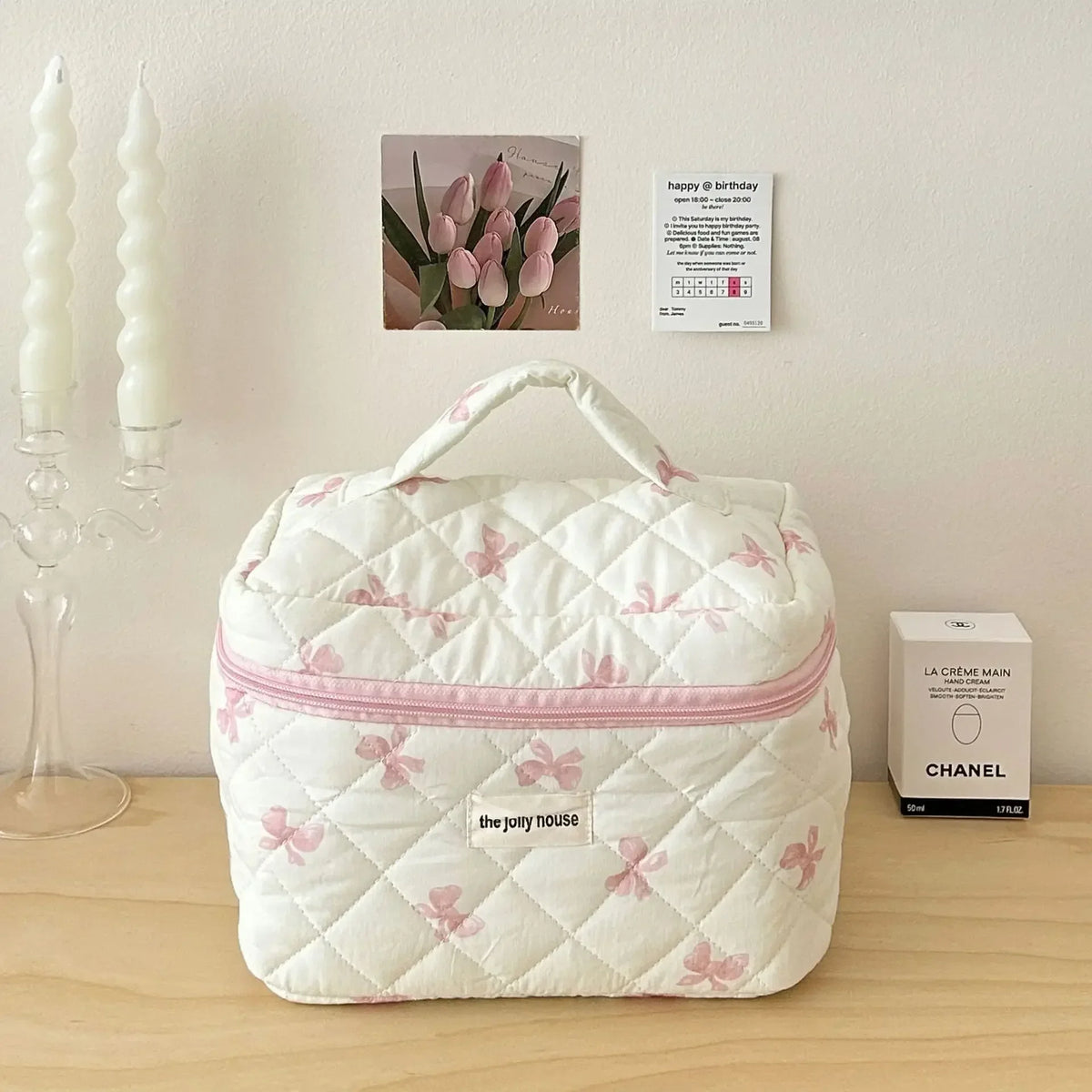 Sac de rangement cosmétique blanc matelassé avec motif nœud rose, posé sur une surface en bois à côté de bougies et d'une boî