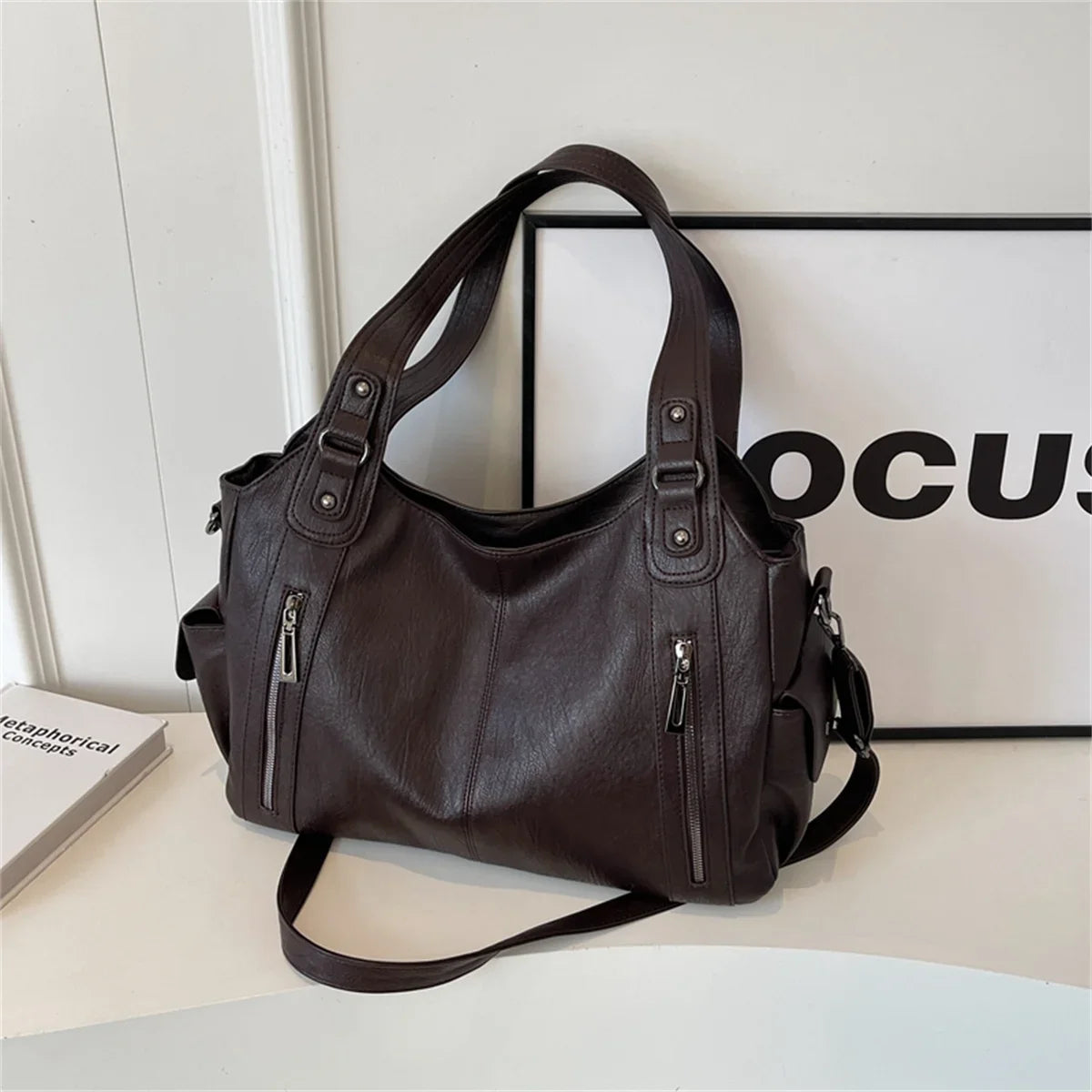 Un sac fourre-tout en cuir marron foncé avec une sangle d'épaule, exposé sur une surface blanche.