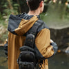 A person wearing a tan jacket and black pants is carrying a tactical chest bag in an outdoor setting.