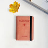 A pink leather passport wallet with a black strap rests on a white surface next to a yellow autumn leaf.
