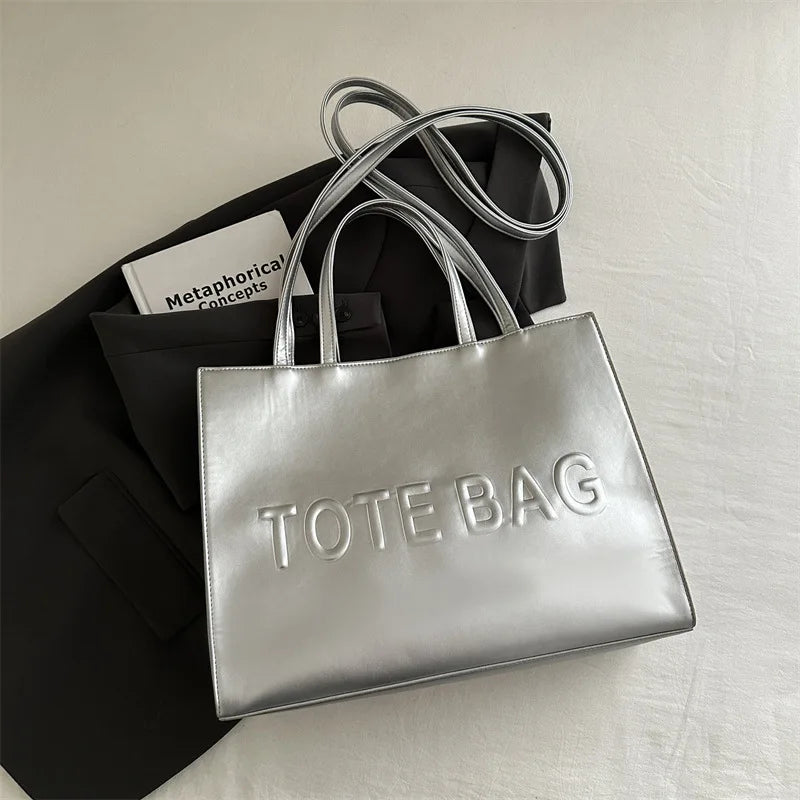 Silver leather tote bag with a long strap resting on a black fabric background.