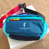 Türkise Outdoor-Sport-Crossbodytasche mit schwarzem Riemen und mehreren Fächern auf einer weißen Box.