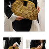 A woman holds a woven straw tote handbag with a natural golden color against a black outfit.