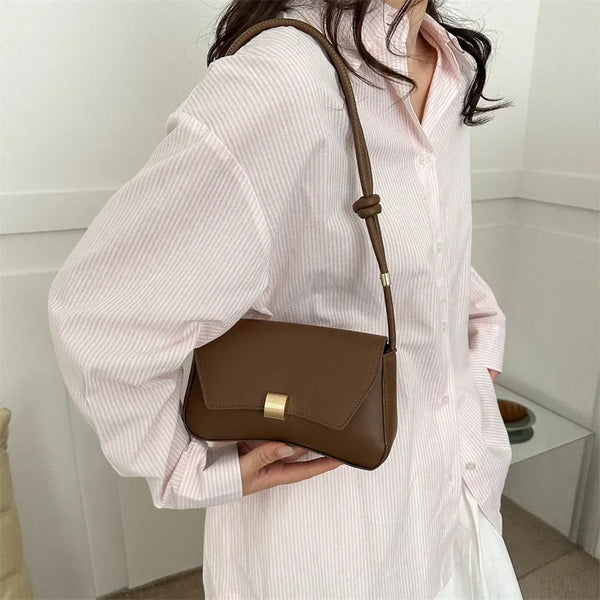 A woman holds a brown leather crossbody bag against a pink striped shirt.
