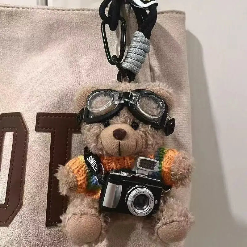 A fluffy bear plush toy keychain featuring a camera and glasses attached to a light beige fabric.