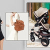 A woman wearing a black dress holds an orange leather backpack and stands next to a large capacity diaper bag with a stroller