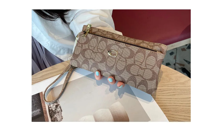 A close-up of a taupe patterned zippered wallet with gold hardware being held by a hand on a wooden surface.