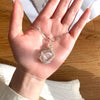 A hand holds a handcrafted amethyst crystal pendant necklace featuring a clear quartz stone wrapped with copper wire.