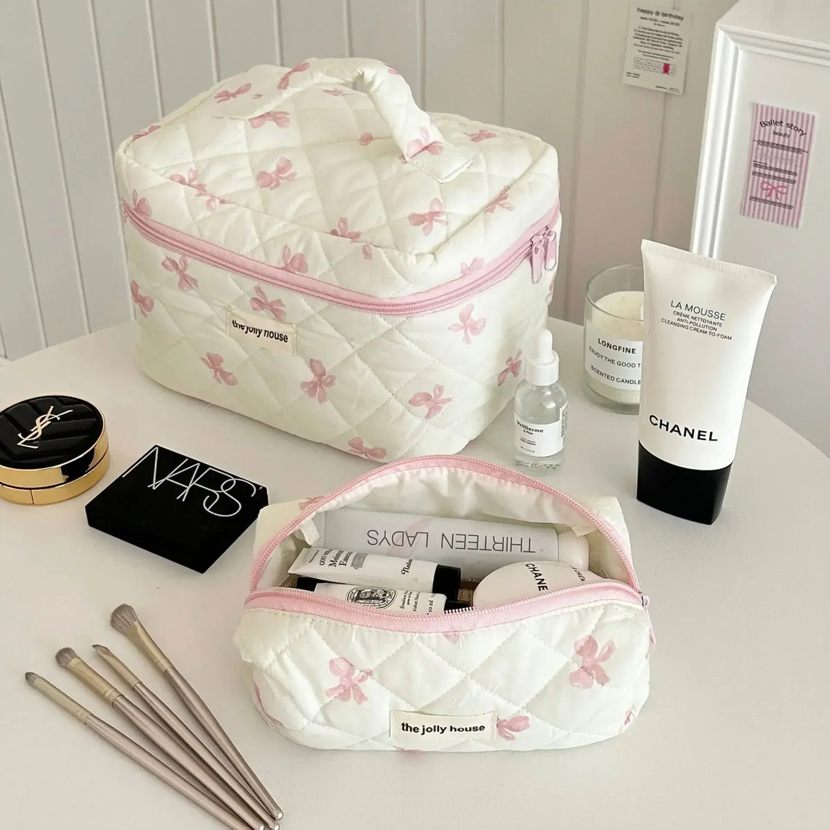 Sac de rangement cosmétique rembourré blanc avec motif de nœud rose, posé sur une surface blanche à côté de produits Chanel e