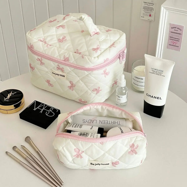 Sac de rangement cosmétique rembourré blanc avec motif de nœud rose, posé sur une surface blanche à côté de produits Chanel e
