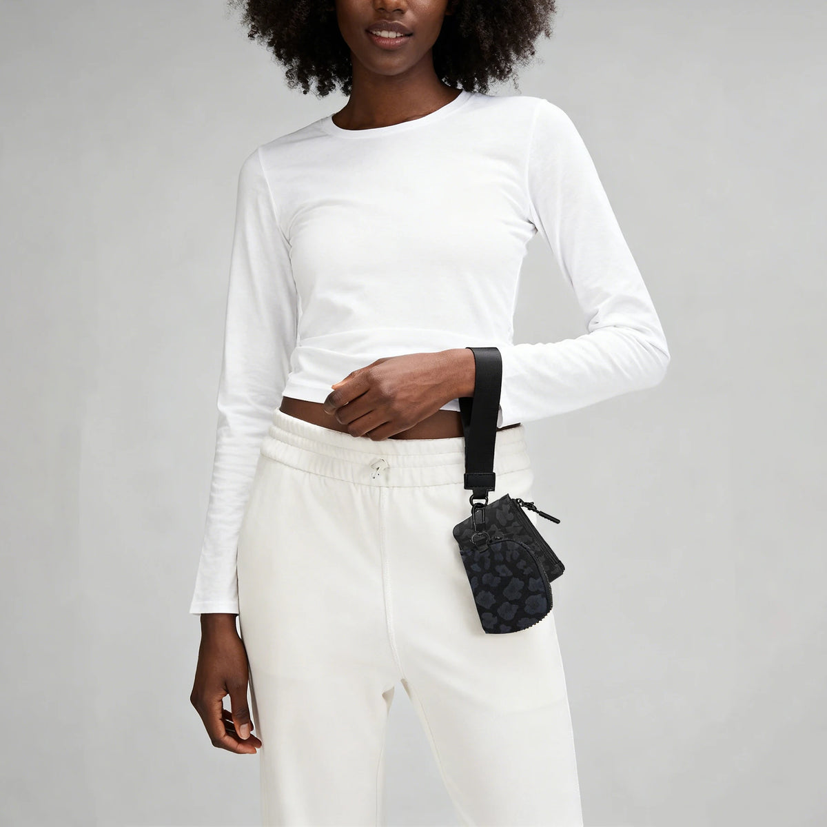 A woman wearing a white long-sleeve top and pants, holding a black nylon wristlet pouch.