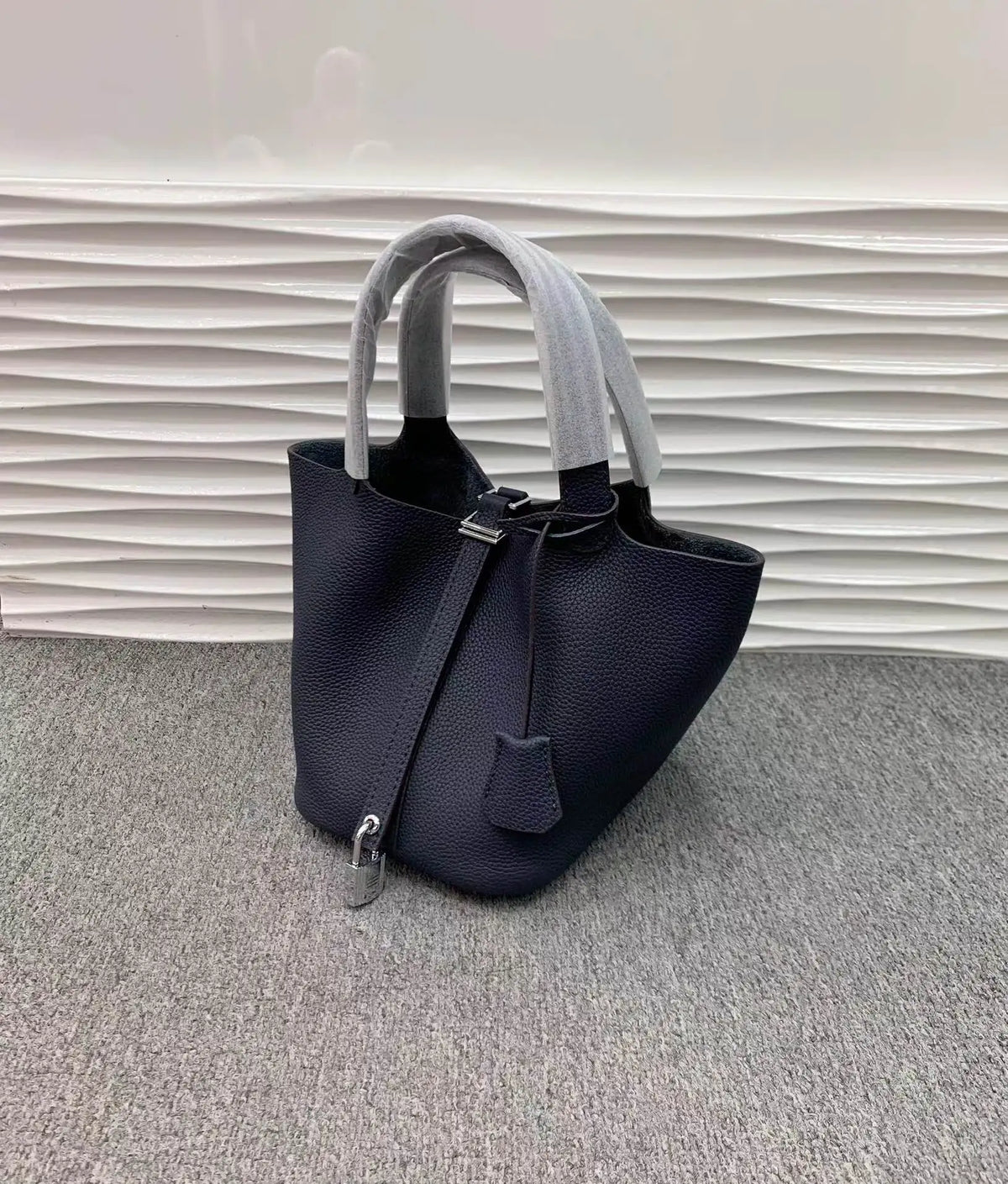 Dunkelblaue Leder-Bucketbag mit hellgrauen Henkeln auf grauem Teppich.