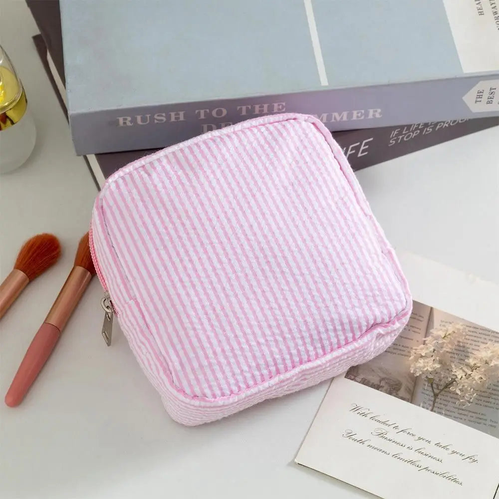 A pink striped storage pouch resting on a white surface next to a book and makeup brush.