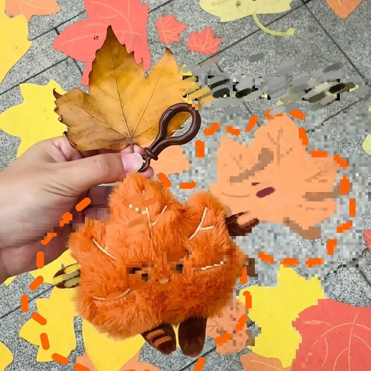 An orange maple leaf plush keychain charm is being held against a background of colorful autumn leaves.
