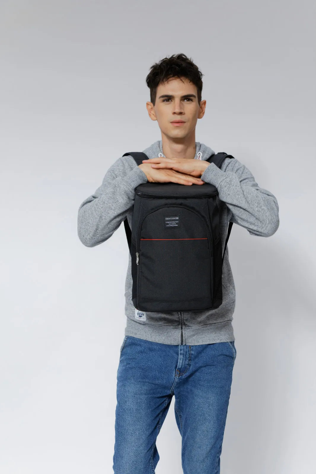 A young man wearing a grey sweatshirt and jeans is holding an Insulated Thermal Cooler Backpack on his shoulders.