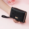 A black compact PU leather women's wallet with a zipper and a leather strap is held in a hand against a pink background.