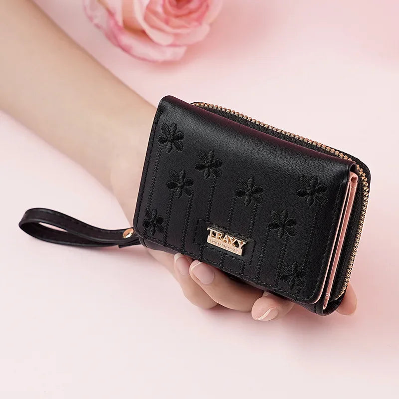A black compact PU leather women's wallet with a zipper and a leather strap is held in a hand against a pink background.