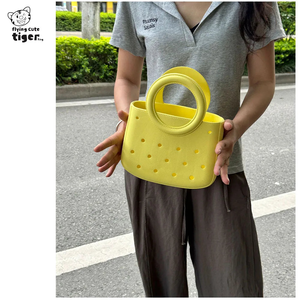A person holds a bright yellow round PVC handbag with a DIY hole design against a blurred outdoor background.