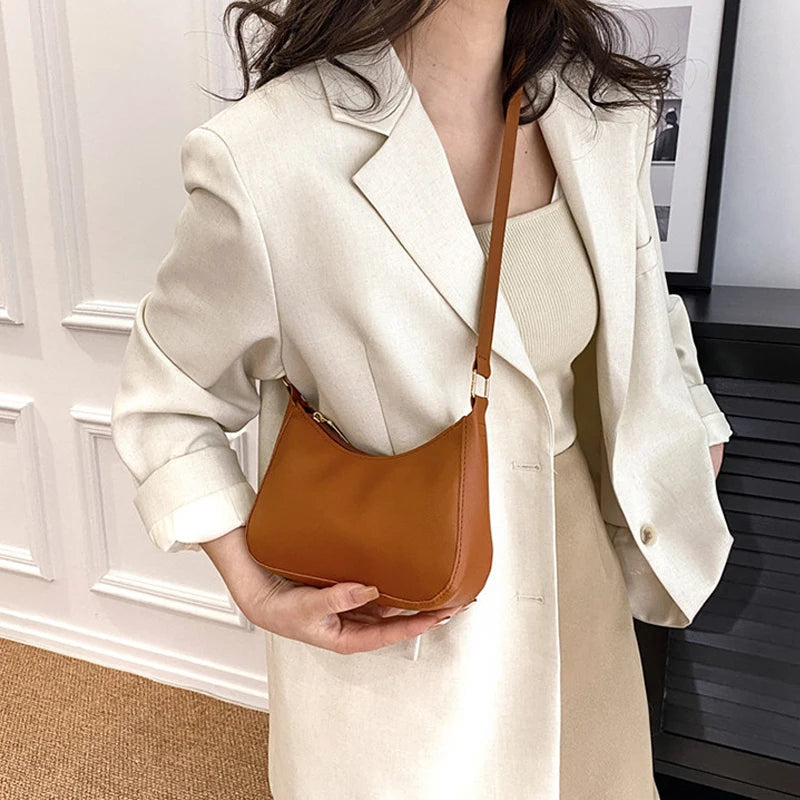 A woman holds a tan leather shoulder bag with a brown strap against a light beige blazer.