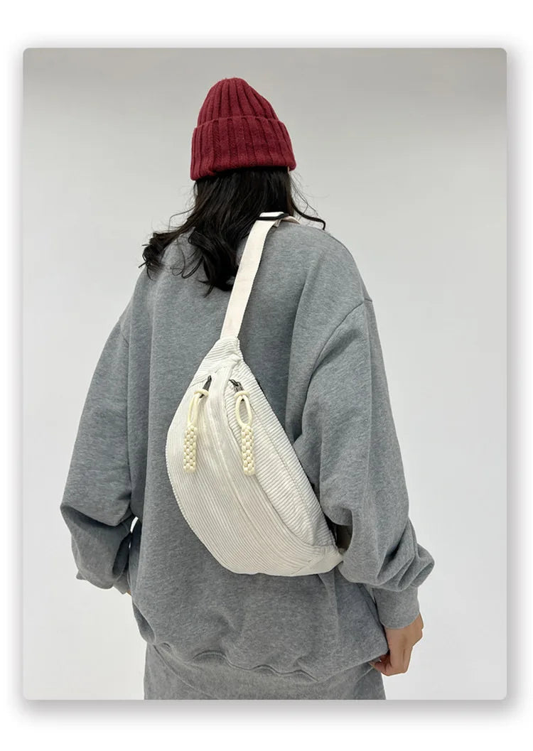 A woman wearing a gray sweatshirt and a maroon beanie is carrying a cream corduroy waist pack.