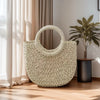 A light green handwoven rattan beach bag hanging against a sheer curtain in a bright room.