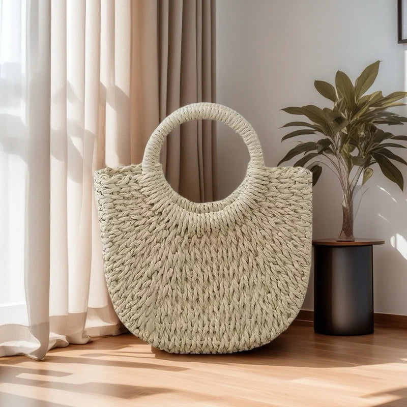 A light green handwoven rattan beach bag hanging against a sheer curtain in a bright room.
