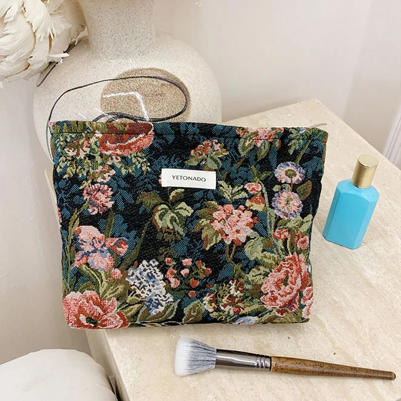 A dark floral patterned cosmetic pouch with pink and green accents rests on a light surface next to a makeup brush.