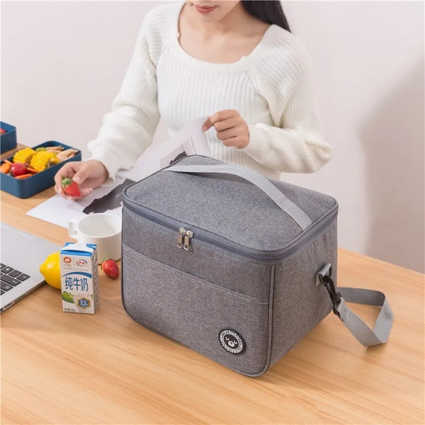 A woman holds a gray insulated thermal lunch bag on a wooden table next to some fruit and a laptop.