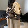 A woman is holding a brown leather tote bag with a strap against a light beige coat.