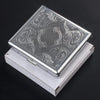 A silver decorative metal cigarette storage box with intricate swirling patterns resting in its white presentation case.