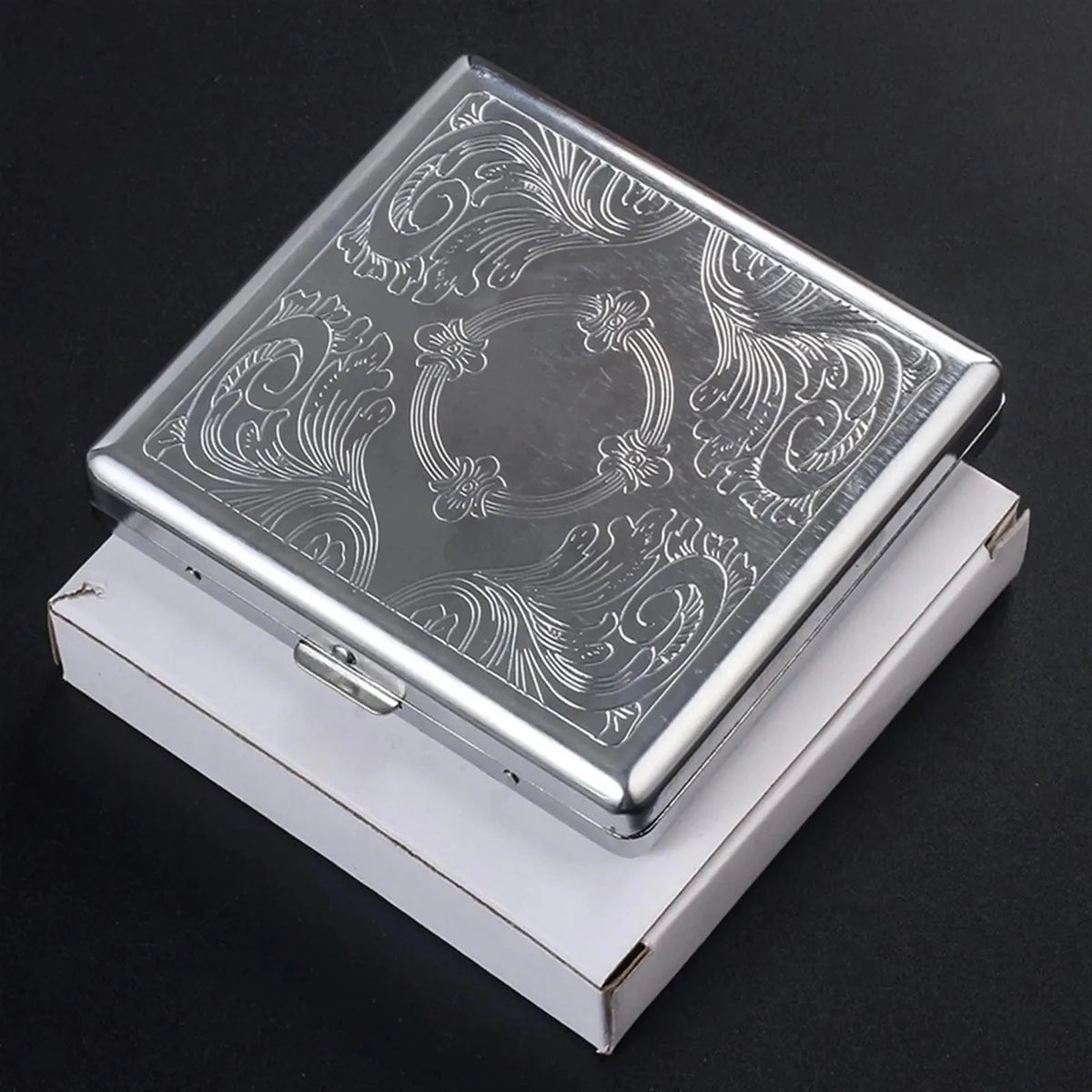 A silver decorative metal cigarette storage box with intricate swirling patterns resting in its white presentation case.
