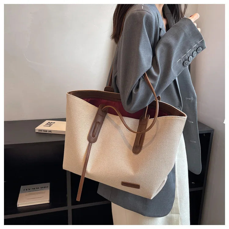 A woman holds a large beige canvas shoulder tote bag with brown leather accents against a light background.