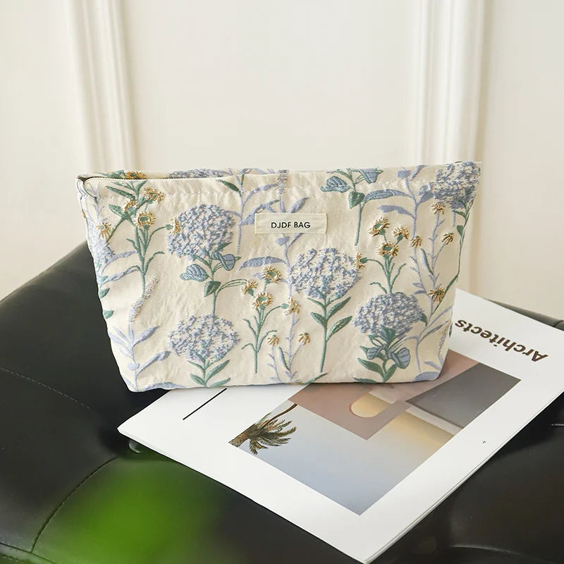A light beige floral canvas makeup bag featuring blue and green botanical patterns, resting on a dark surface.