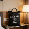 A black laundry bag with a handle is placed on a metal rack next to a bedside lamp.