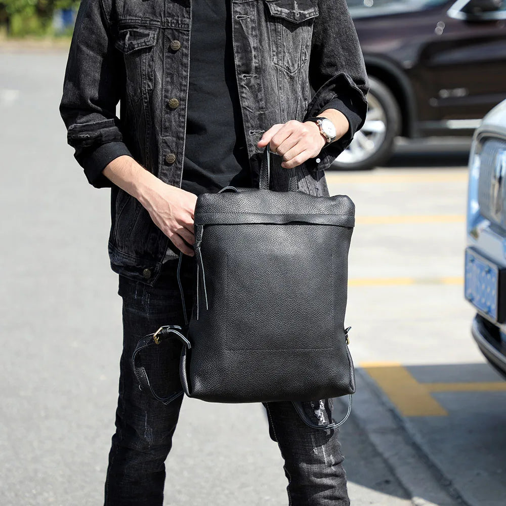 A person is holding a black full-grain leather backpack in an outdoor setting.