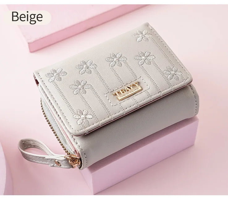 A beige compact PU leather women's wallet with a zipper closure and floral embroidery is shown resting on a pink surface.