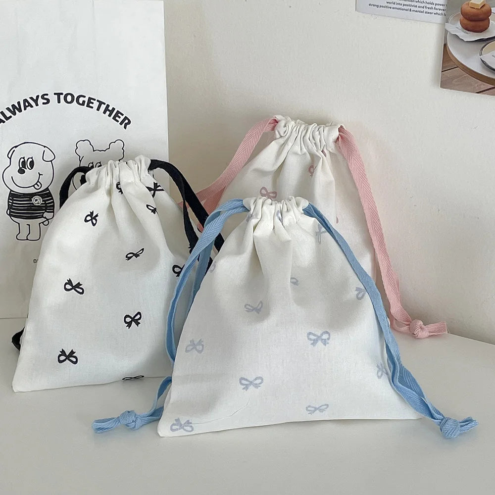 Three canvas makeup bags in white, pink, and blue with bowknot ties are displayed on a white surface.