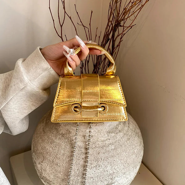A hand holds a gold square crossbody bag with a chain strap resting on a fuzzy gray spherical object.