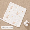 A white muslin baby burp cloth towel with small bunny embroidery is shown on a beige textured surface.