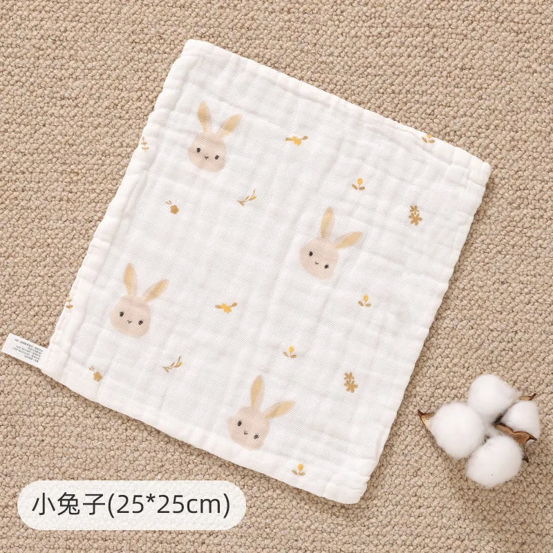A white muslin baby burp cloth towel with small bunny embroidery is shown on a beige textured surface.