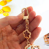A hand holds a gold-colored flower bag extender chain with a clasp and decorative charms.