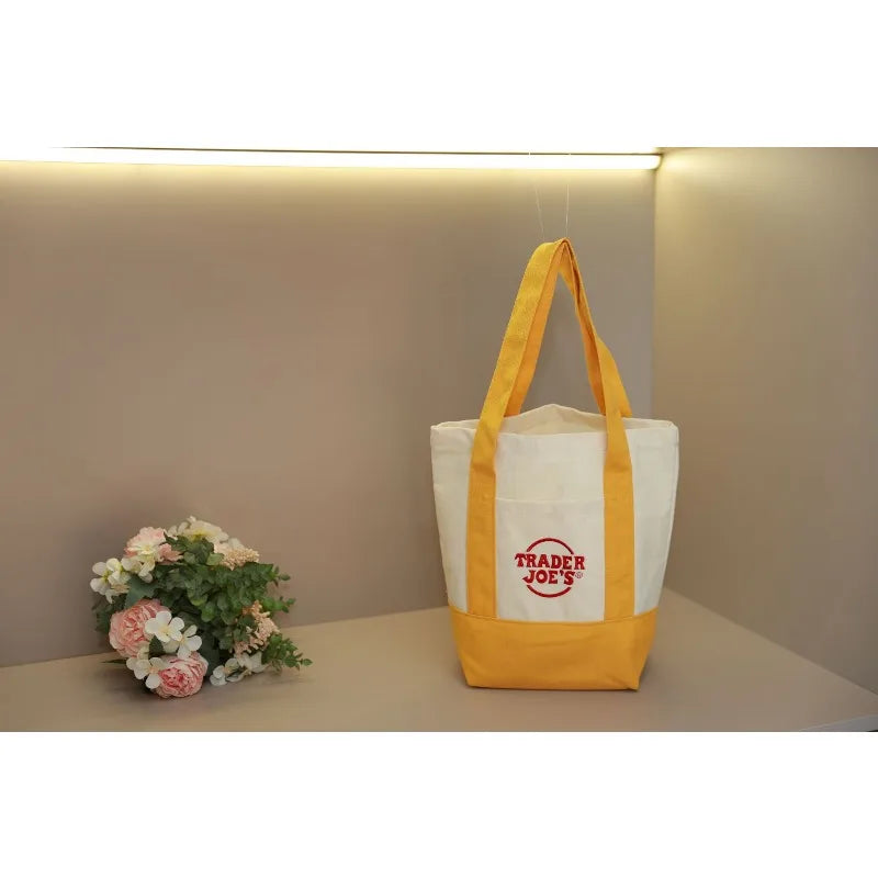 A yellow canvas tote bag with white stripes and a red logo hanging against a light gray wall.