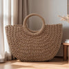 A large, woven natural fiber beach bag with a handle is displayed on a wooden floor next to a window.