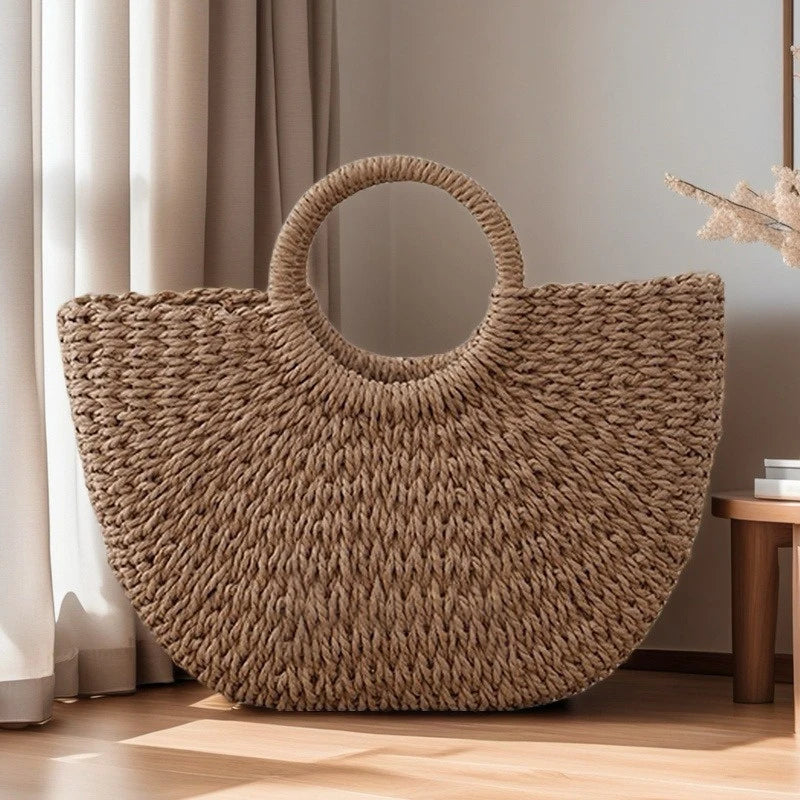 A large, woven natural fiber beach bag with a handle is displayed on a wooden floor next to a window.