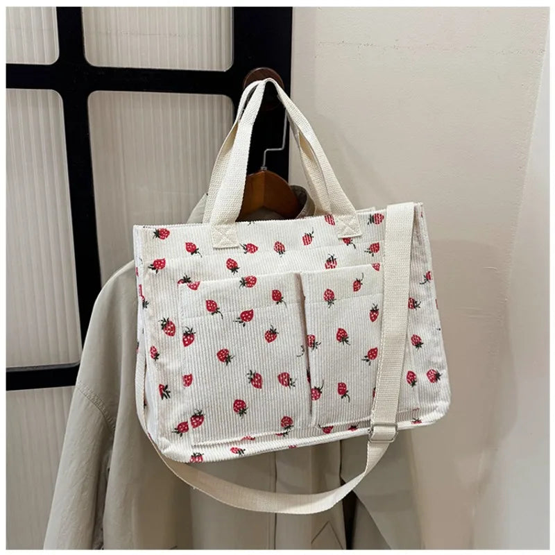 A white canvas tote bag featuring a pattern of red strawberries on a light background, displayed on a wooden hanger.