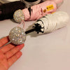 A hand holds a compact rhinestone umbrella with a sparkly sphere and white fabric, next to a pink fabric roll.