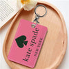 A pink tag with black text spelling out Kate Spade New York is attached to a silver keychain on a wooden surface.