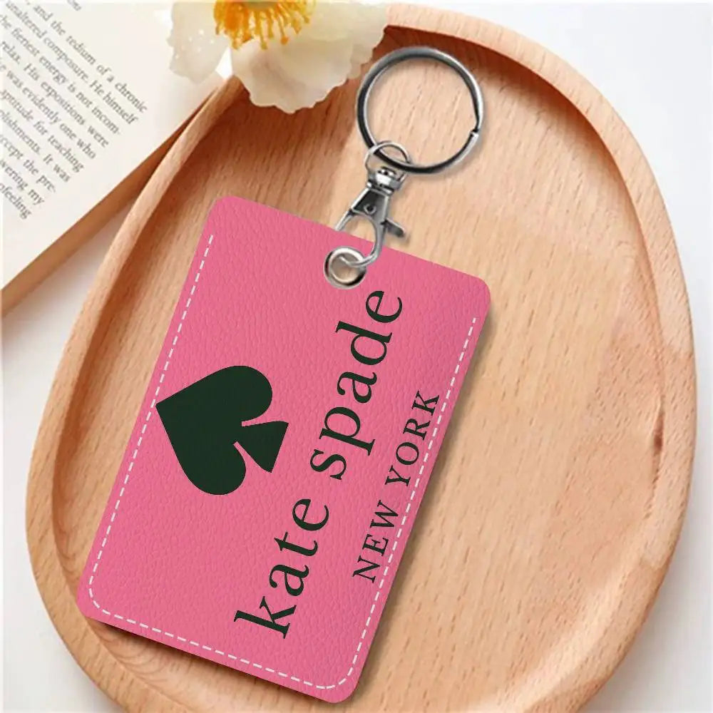 A pink tag with black text spelling out Kate Spade New York is attached to a silver keychain on a wooden surface.