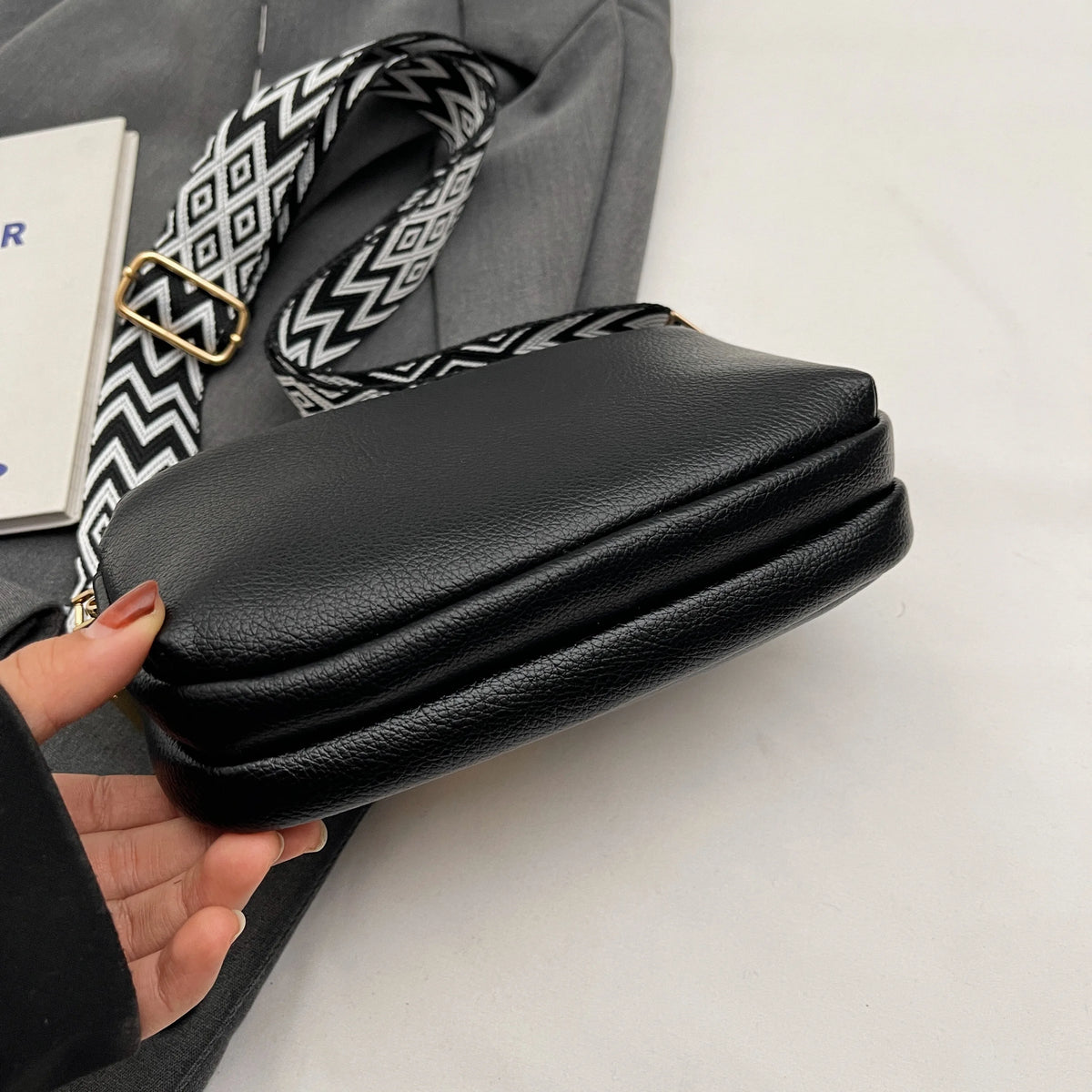 A black vegan PU crossbody bag with a patterned strap is shown next to a gray garment.