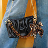 Schwarze Kunstleder-Tasche mit Leopardenmuster-Riemen und Schleifchen-Anhänger an blauen Jeans.